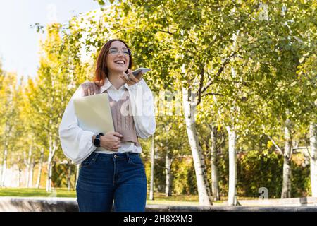 Positive smiling businesswoman with glasses recording audio message or using voice recognition, holding laptop while walking in the park Stock Photo