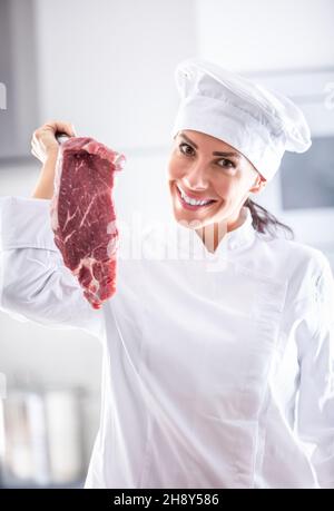 Female chef in professional white clothing holds a big chunk of beef steak in her hand, smiling. Stock Photo