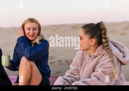 Two blonde women talking while drinking tea sitting on a beach Stock Photo