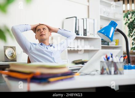 Relaxed office worker sitting at workplace with hands behind head Stock Photo