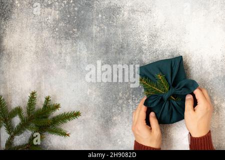 Furoshiki-style gift and spruce for Christmas or New Year on a grey background with hands. Flat lay. Copy space. Stock Photo