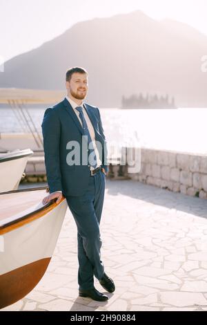 Groom in a blue suit stands leaning on the stern of the boat on the coast Stock Photo