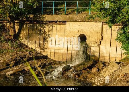 Wastewater is discharged into the river. Dirty water flows from a pipe in the concrete wall. The concept of ecology Stock Photo