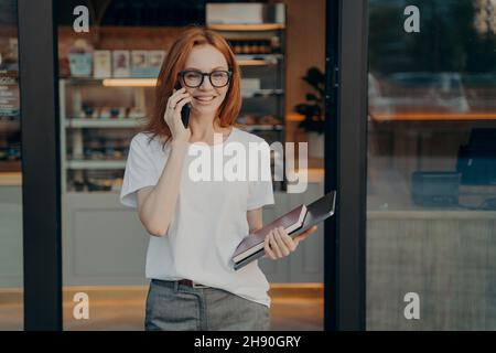 Smiling woman with laptop and notebook in hand enjoying talk on cellphone near coffee shop Stock Photo