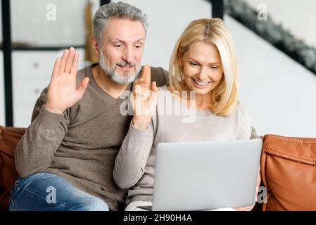 Lovely mature spouses using laptop computer for online communication, video connection with friends or family on the distance. Happy middle-aged couple sits on sofa, looks at the webcam and waving Stock Photo