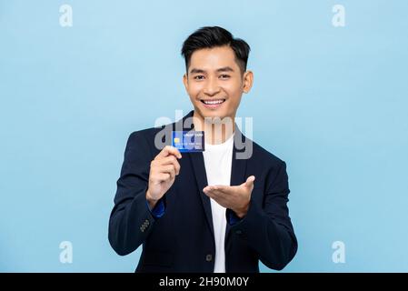 Handsome smiling Asian man in semi formal suit showing credit card in light blue isolated studio background Stock Photo