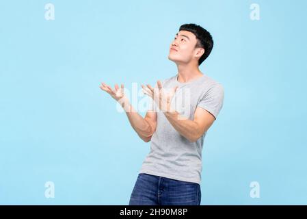 Asian man raising open hands with shocked face and looking at empty space above isolated on studio light blue background Stock Photo