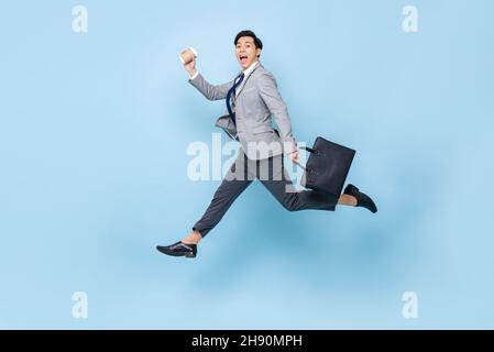 Energetic young handsome Asian businessman jumping in mid air with bag and coffee cup in isolated light blue studio background Stock Photo