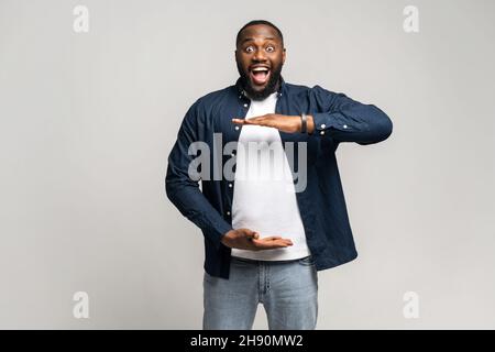 Excited surprised African man holding hands in front of him, presenting something, shocked dark skinned guy holding empty space for your product isolated on gray, demonstrates size. Mockup concept Stock Photo