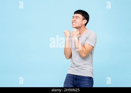 Happy ecstatic young Asian man raising his fists doing yes gesture celebrating success in isolated light blue studio background Stock Photo