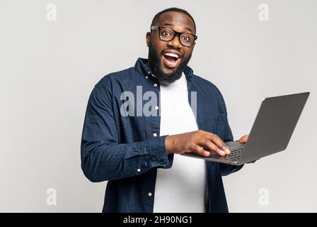 Excited surprised black man standing with laptop on grey background, typing, feeling stunned by online news looking at camera, trader investor surprised by stock market changes Stock Photo
