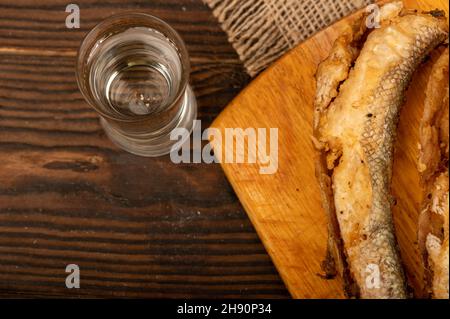 A small fried fish on a wooden board, pieces of bread and a glass of vodka, close-up, selective focus Stock Photo