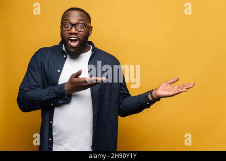 Amazed African American man in glasses standing isolated over yellow background pointing with palms on the blank space for advertisement, mockup image, check this out, funny concept Stock Photo