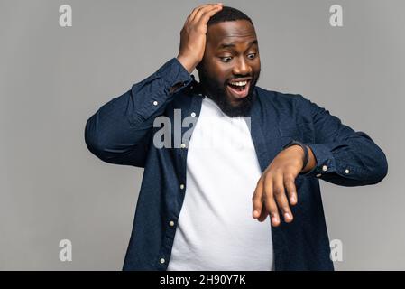 Surprised African-American man wearing casual shirt over t-shirt is late, looks at the hand watch and holding amazed, male office employee forgot about meeting, isolated on grey Stock Photo