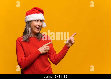 Positive elderly woman in red santa hat looks and points to the side with both index fingers, isolated over yellow background, copy space, advertises or recommends a new product, holiday sale Stock Photo