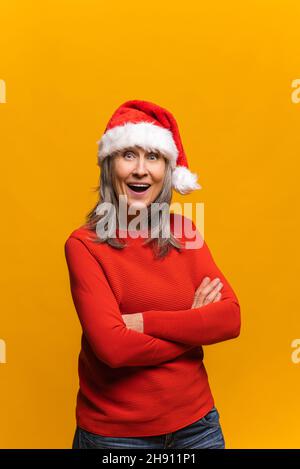 Joyful senior gray-haired woman in santa hat stands with arms crossed and looks at the camera, positive mature middle-aged lady smiles, celebrating new year and christmas isolated on yellow background Stock Photo