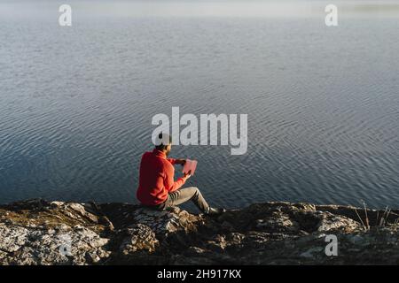 Man reading book while sitting on rock at lakeshore Stock Photo