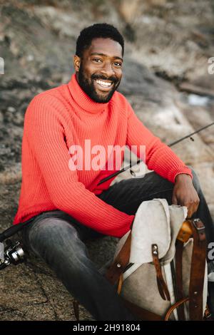Smiling man sitting with backpack and fishing rod on rock Stock Photo