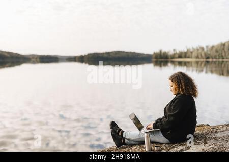Woman working on laptop remotely at lakeshore during weekend Stock Photo