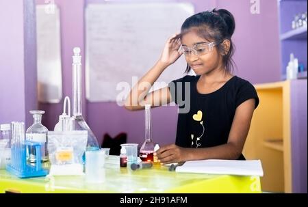 Confused girl kid scratching head by looking lab equipments at chemistry laboratory while making experiment - concept of difficulty in learning at Stock Photo