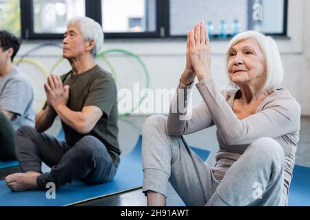 Grey haired woman practicing yoga near blurred asian friend in sports center Stock Photo