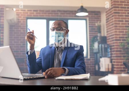 Entrepreneur thinking about business strategy and using laptop during covid 19 pandemic in office. Pensive man looking at computer while he thinks about financial solution, wearing face mask. Stock Photo