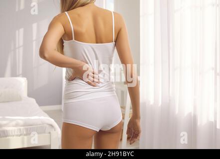 Woman having backache or kidney pain standing in bedroom and holding hand on lower back Stock Photo