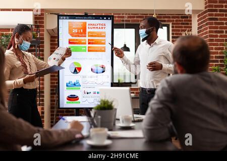 Businessteam with medical face mask to prevent infection with covid19 showing company graphs discussing business strategy in startup office. Diverse people working at financial presentation Stock Photo