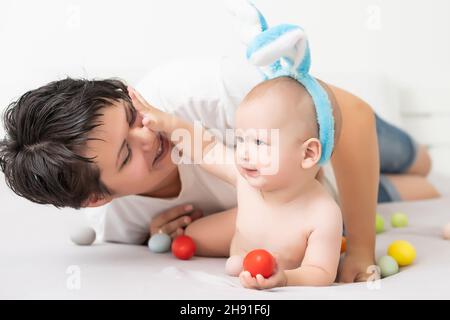 Mom and baby with rabbit ears, with Easter eggs in their hands, sitting on the sofa, parents and children play indoors. Family celebrates Easter Stock Photo