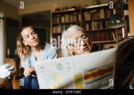 Senior woman reading reading newspaper by female healthcare worker in living room Stock Photo