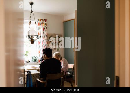 Young male healthcare worker drinking coffee with senior woman in kitchen at home Stock Photo