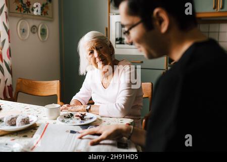 Young male nurse reading newspaper while elderly woman looking at him in kitchen Stock Photo