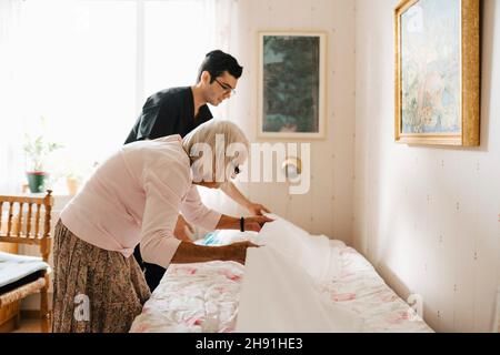 Senior woman and male healthcare worker placing blanket on bed at home Stock Photo