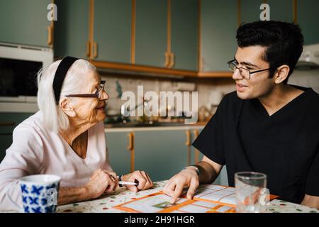 Smiling senior woman talking with male nurse in kitchen at home Stock Photo