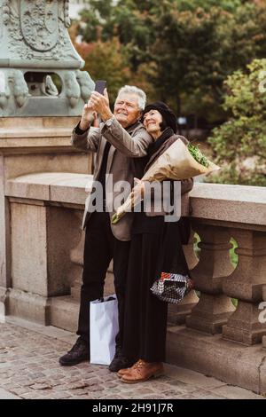 Happy senior couple taking selfie through smart phone while standing on bridge Stock Photo