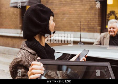 Senior woman with smart phone looking at man while entering car in city Stock Photo
