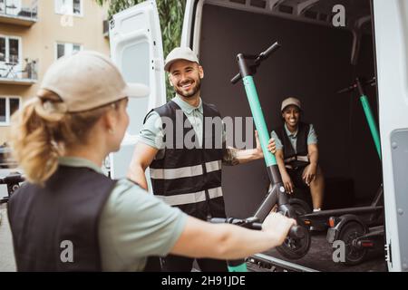 Smiling male worker talking with female coworker while loading push scooter in delivery van Stock Photo