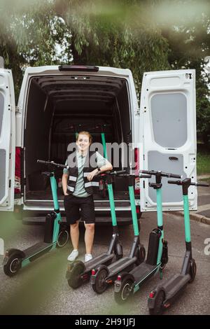 Young delivery woman with hand in pocket leaning on electric push scooter against van Stock Photo
