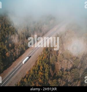 Aerial shot of a lorry truck drives on an intercity road surrounded with autumn forest and with clouds above. Fall landscape Stock Photo