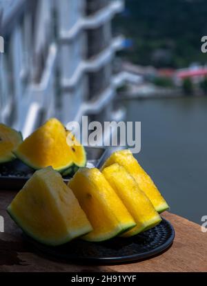 Fresh juicy yellow watermelon cut into triangles on round plate and background of buildings and city view. Tropical fruits. Select focus. Stock Photo