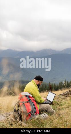 a bearded Caucasian man with a laptop sits on a fallen tree in the mountains with a beautiful view of the valley. Stock Photo