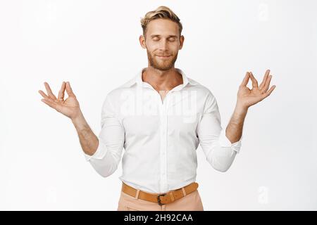 Smiling relaxed blond man meditating with closed eyes, feeling at ease during meditation, resting his mind and breathing freely, standing over white Stock Photo