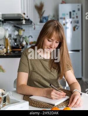 Young female making notes in notebook sitting in kitchen. Planning buying. Making list of products Stock Photo