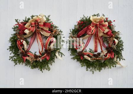 A pair of natural evergreen holiday wreaths with pine cones, winterberries, nuts, cinnamon sticks and ribbons on an old rustic wood sided barn with pe
