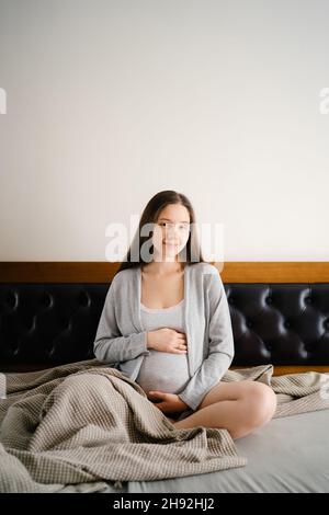 Pregnant woman sits on the bed and hugs her belly with her hands Stock Photo