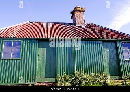 Looking up at old moorland bothy in Highland Perthshire with stone chimney, rusted corrugated iron roof and green painted wooden walls and doors. Stock Photo