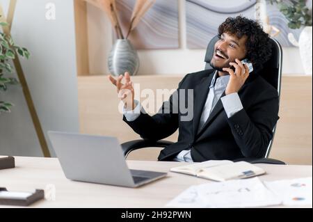 Positive successful Indian businessman, IT specialist, marketolog or manager, sitting at work desk in the office, having a pleasure cell phone conversation during work time, smile friendly, looks away Stock Photo