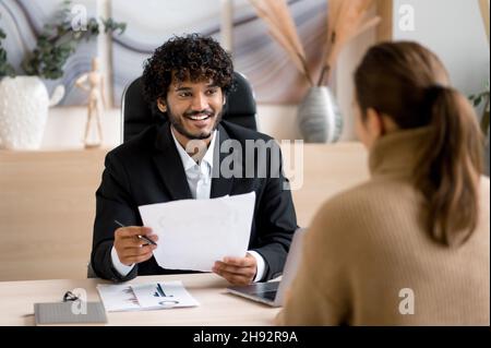 Confident successful Indian male HR manager, entrepreneur, conducts interview girl in a modern office, wearing formal clothes, looking and listening to a job candidate, holding a resume in hands Stock Photo