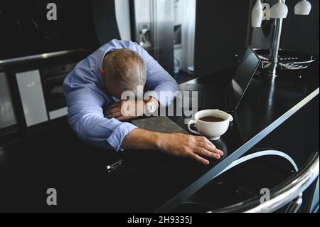 Tired mature businessman, entrepreneur asleep at work table with laptop and half drunk cup of coffee. High angle view Stock Photo