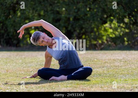 A middle aged woman practicing yoga barefoot outside in a grassy park. She is wearing a grey and blue vest and black leggings. The style of yoga she i Stock Photo